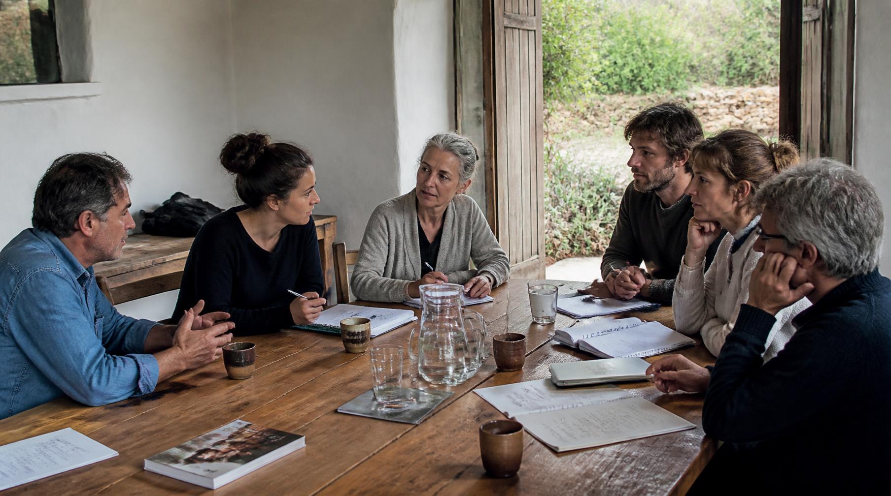 Pequeno grupo de adultos de idades diferentes à volta de uma mesa comprida de madeira, numa sala caiada com portas abertas para o jardim.