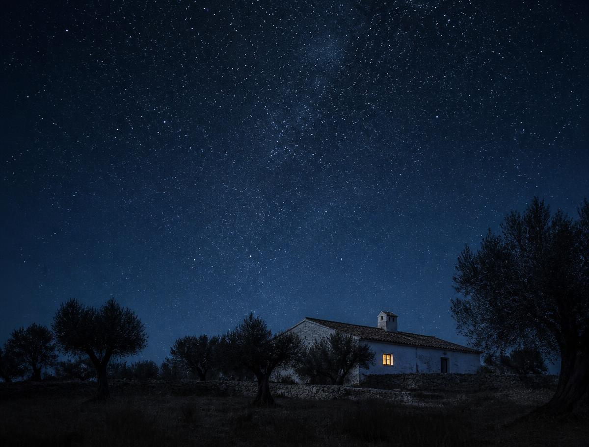 Casa baixa caiada sob um céu nocturno cheio de estrelas, com uma janela iluminada.