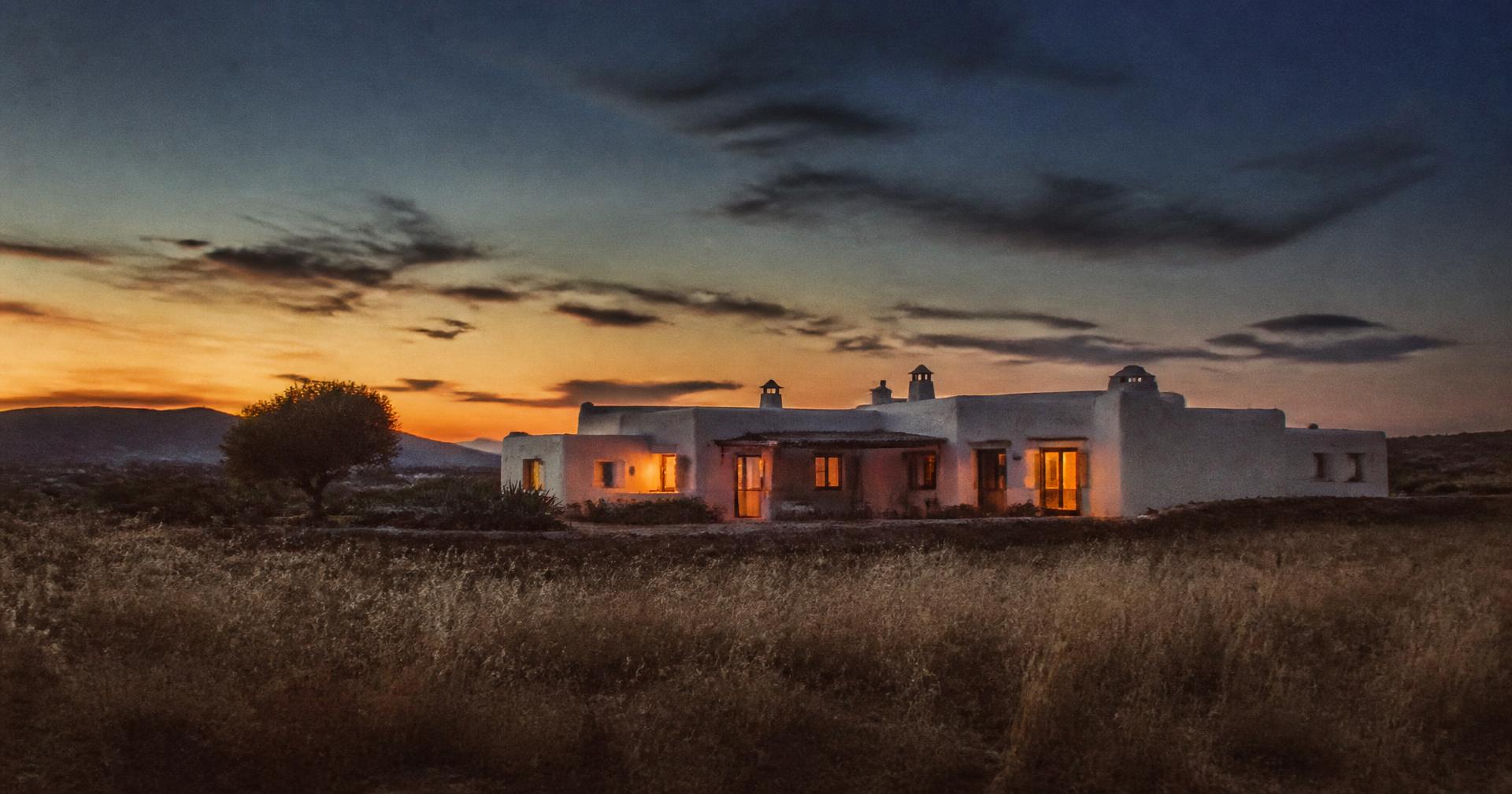Casa caiada ao anoitecer, vista através de um campo de ervas secas, com as janelas iluminadas.